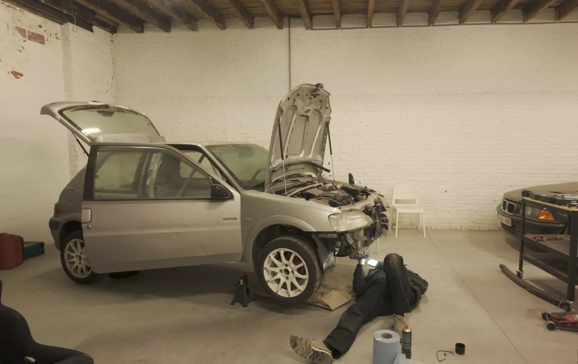 The car on jack stands with its hood open and front bumper removed, a mechanic working underneath
