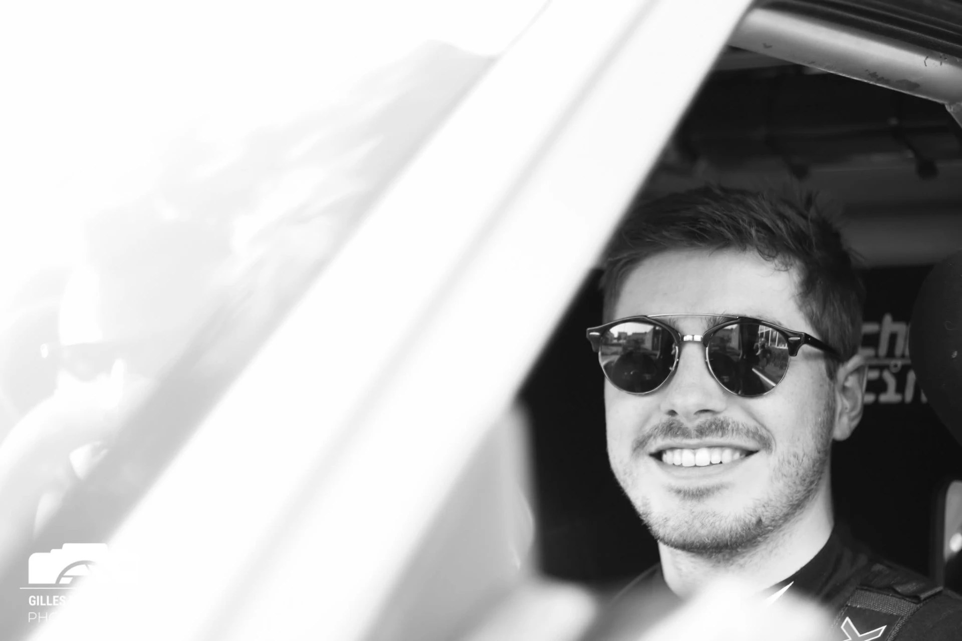 Louis Van de Vyvere smiling in the cockpit, wearing sunglasses and a racing harness, in black and white