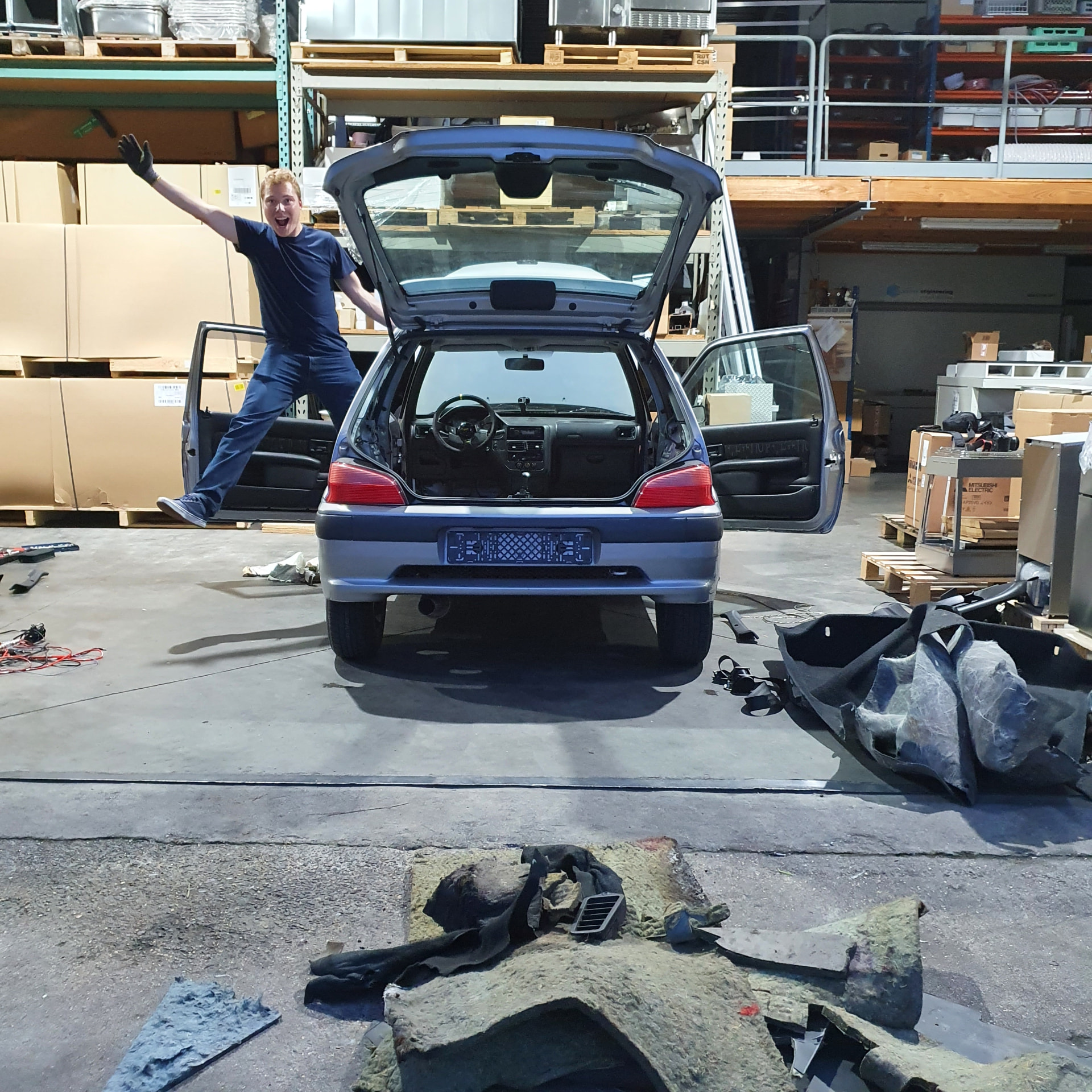 The car with its doors and hatch open in a warehouse, interior stripped, one person jumping in the doorway